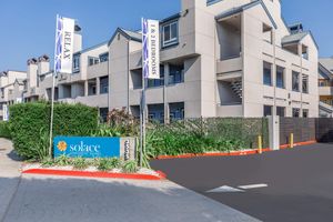 A residential apartment community named "Solace" featuring a welcoming sign displaying "1 & 2 Bedrooms." The entrance is landscaped with greenery, and flags are visible providing directions to relax. The building exterior is light-colored, with multiple stories and a modern design.