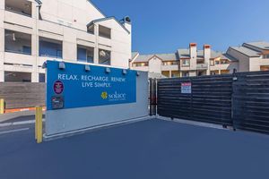 A gated entrance to Solace Apartment Homes featuring a blue sign with the text “RELAX. RECHARGE. RENEW. LIVE SIMPLY.” The background shows two multi-story apartment buildings with colorful accents. The area is clean and well-maintained, indicating a welcoming residential environment.