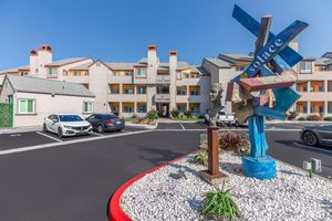 A colorful abstract sculpture titled "Solace" stands in front of a three-story residential building with a modern design. Nearby, parked cars are visible on an asphalt lot, surrounded by landscaped areas featuring white gravel and green plants under a clear blue sky.