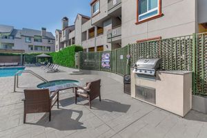 Outdoor pool area featuring a swimming pool, hot tub, barbecue grill, and seating with two chairs. Surrounding greenery and residential buildings create a relaxed atmosphere. Clear blue skies enhance the serene setting, perfect for leisure and relaxation.