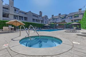 A sunny outdoor pool area featuring two swimming pools, a hot tub, and several lounge chairs. Surrounding greenery offers privacy, and shaded seating is available under a large umbrella. The scene conveys a relaxed atmosphere suitable for leisure and enjoyment.