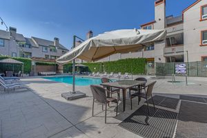 A sunny poolside area featuring a clear blue pool, surrounded by lounge chairs and a shaded table with chairs under a large umbrella. Green hedges provide privacy, and a multi-story building can be seen in the background. The scene is inviting for relaxation and outdoor enjoyment.