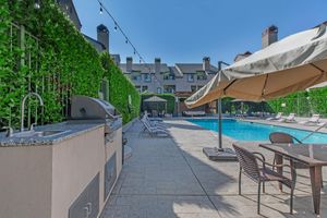 A spacious pool area featuring a glistening blue swimming pool surrounded by lounge chairs. There is a shaded cabana and a barbecue grill station nearby, all set against a backdrop of well-maintained greenery and a clear blue sky. Ideal for relaxing and socializing outdoors.