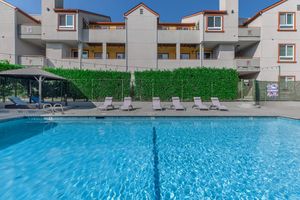 A modern pool area with lounge chairs arranged around a clear blue swimming pool. In the background, a multi-story building features balconies and greenery. The scene is well-lit with a clear blue sky, creating a relaxing atmosphere for leisure and enjoyment.