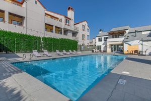 A clear blue swimming pool surrounded by lounge chairs and lush greenery, located near a modern building. The sky is bright and sunny, enhancing the inviting atmosphere of the pool area.