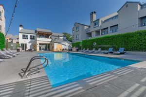 A clear blue swimming pool surrounded by lounge chairs, with a modern building structure in the background. Lush green hedges line the area, and the sky is bright and sunny, creating a relaxing outdoor atmosphere.