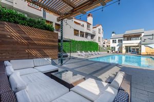 A modern outdoor pool area featuring a spacious lounge with white cushions, a glass-topped table, and wooden privacy screens. Sun loungers are arranged beside a sparkling blue pool, with well-maintained greenery in the background and bright blue skies above, creating a relaxing atmosphere.