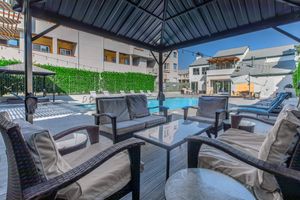 A shaded outdoor seating area with comfortable wicker chairs and a glass table overlooks a clear swimming pool. The pool area is surrounded by green hedges and modern buildings, creating a relaxing atmosphere. Sun loungers are placed nearby for leisure.