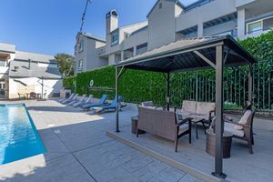 A sunny poolside area featuring a swimming pool, comfortable lounge chairs, and a shaded gazebo with wicker seating. Lush greenery lines the background, providing a serene atmosphere for relaxation and leisure. The setting is ideal for outdoor enjoyment and social gatherings.