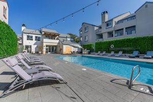 A modern outdoor swimming pool area with lounge chairs arranged along the sides. The pool is surrounded by green foliage and an apartment building. There are decorative string lights above the pool, and a few umbrellas for shade are visible. The sky is clear and blue, creating a bright, inviting atmosphere.