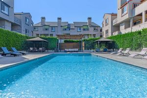 A clear blue swimming pool surrounded by lounge chairs and shaded gazebos. In the background, there are modern apartment buildings with greenery along the perimeter. The sky is bright and sunny, creating a relaxing outdoor atmosphere.