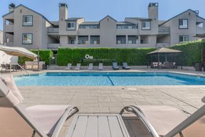 A tranquil pool area featuring a clear blue swimming pool surrounded by lounge chairs, with lush greenery and a modern building in the background. The scene is sunny and inviting, perfect for relaxation and leisure.