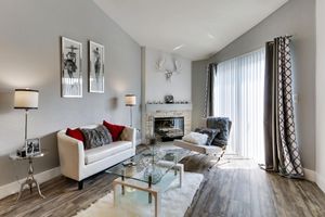 A modern living room featuring a white sofa with red and gray accent pillows, a glass coffee table, and a plush area rug. There are framed wall art pieces, a stylish fireplace, and large windows with sheer curtains. The decor is elegant and minimalist, with neutral tones and a cozy atmosphere.
