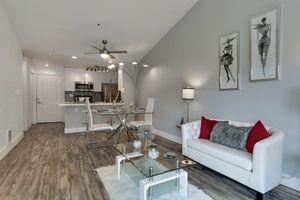 A modern living room featuring a stylish white couch with red and gray throw pillows, a glass coffee table, and decorative items. There’s a dining area in the background with a sleek table and chairs. The walls are painted light gray, and there are framed artworks depicting stylish figures. A ceiling fan adds a cozy touch.