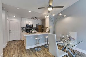 Modern kitchen and dining area featuring a sleek design. The kitchen showcases white cabinets, stainless steel appliances, and a tiled backsplash. A glass dining table is set with elegant tableware, surrounded by white chairs. A contemporary light fixture and fan add a stylish touch to the space. Light wood flooring completes the look.