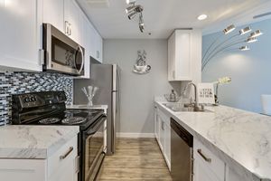 A modern kitchen featuring white cabinets, a marble countertop, and stainless steel appliances. It includes an oven, a refrigerator, and a double sink. The backsplash is made of small black and white tiles. Decorative elements, like a coffee-themed wall art and a plant, add a stylish touch to the space.