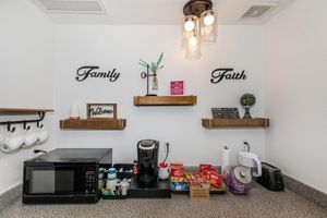 A cozy kitchen corner featuring shelves with decorative signs reading "Family" and "Faith." Below, there's a microwave, a coffee maker, and various snacks in boxes. The space has a welcoming feel, with a small plant and cups hanging on the wall. Warm lighting adds to the inviting atmosphere.