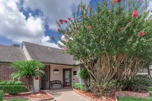 A charming entrance to a building featuring a covered porch, surrounded by lush greenery and flowering trees. The pathway leads to a door, with well-maintained landscaping including palms and colorful flowers, under a blue sky with fluffy white clouds.
