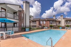 A brightly lit outdoor swimming pool surrounded by a fence, with lounge chairs and tables nearby. In the background, there are two apartment buildings with balconies and a clear blue sky with fluffy white clouds.