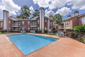 A clear blue swimming pool surrounded by a paved patio in an apartment complex. There are lounge chairs and an umbrella near the pool. The buildings are two stories with brick exteriors and balconies, set amidst trees and greenery under a partly cloudy sky.