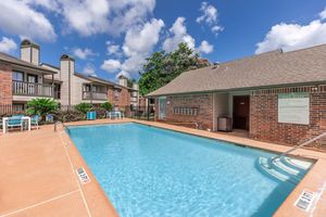 A sunny outdoor pool area surrounded by brick buildings with balconies, featuring lounge chairs and a small pool house. The pool is clear and inviting, with blue skies and fluffy white clouds above. Lush greenery adds a touch of nature to the setting.