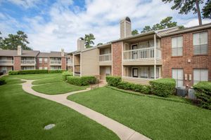 A well-maintained apartment community featuring two buildings with a landscaped grassy area and curved pathways. The buildings have balconies and are surrounded by trees under a partly cloudy sky.