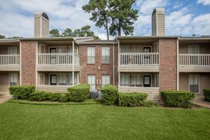 Two-story brick apartment building with balconies, surrounded by neatly trimmed bushes and a green lawn under a partly cloudy sky.