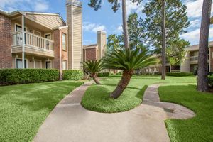 A well-maintained apartment community featuring green lawns and palm trees, with a walkway curving through the landscaping and residential buildings in the background.