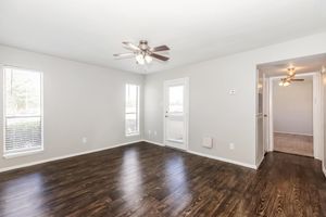 A spacious and well-lit living room featuring hardwood floors, a ceiling fan, and multiple windows. The walls are painted in a light color, and there is a door leading to the outside and a hallway to another room. The overall atmosphere is bright and inviting.