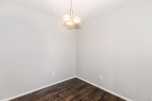 A well-lit empty corner of a room featuring a simple chandelier hanging from the ceiling, light gray walls, and dark wood flooring. The space appears clean and uncluttered, suitable for various uses or decoration.