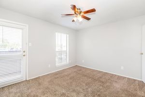 A well-lit room featuring light gray walls and plush beige carpeting. There is a ceiling fan with wooden blades, and two windows with white blinds. A door leads outside, and the overall space appears clean and inviting, suitable for various uses.