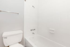 A clean, simple bathroom featuring a white bathtub, a showerhead, and a toilet. The walls and fixtures are white, creating a bright and airy atmosphere. There is a towel bar mounted on the wall, and the overall design is minimalistic.