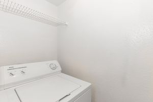 A clean, bright laundry room featuring a white washing machine against a plain white wall. A wire shelf above provides storage space, enhancing the functional and minimalist design of the area. The room appears well-maintained and organized.