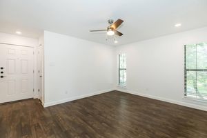 Bright interior of a room featuring light wooden flooring, a ceiling fan, and two windows with blinds. The walls are painted white, and a door is visible on the left side. The space is empty and well-lit, providing a sense of openness and potential for decoration.