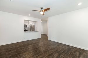 A spacious and bright living area with wood-like flooring, a ceiling fan, and a modern design. The room features white walls and a view of a kitchen area with stainless steel appliances and a granite countertop, creating an open and inviting atmosphere.