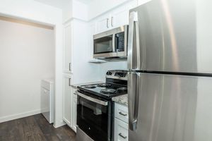 A modern kitchen showcasing stainless steel appliances including a refrigerator, microwave, and stove. The cabinets are white with a granite countertop. A washer and dryer are partially visible in an adjacent space, and the floor is dark wood. Overall, the design is clean and contemporary.