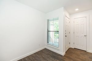 A bright and spacious room featuring white walls, wooden flooring, and a window with blinds. There is a doorway leading to another room and a closet door visible. The area is well-lit and has a clean, modern aesthetic.