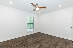 A bright, empty room featuring a ceiling fan, white walls, and a large window with blinds. The floor is made of dark wood, creating a modern and clean aesthetic. Natural light filters in from the window, enhancing the spacious feel of the room.
