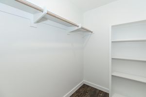 A clean, empty closet featuring a white wall and a wooden rod for hanging clothes. On one side, there is a simple white shelf unit, while the flooring has a dark, wooden finish. The overall ambiance is bright and spacious, emphasizing minimalism and organization.