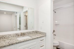 A modern bathroom featuring a white vanity with a granite countertop, a large mirror above the sink, and a shower area with a bathtub. The walls are painted white, and there are bright overhead lights, creating a clean and spacious appearance.
