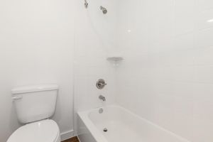 A clean, modern bathroom featuring a white bathtub with a showerhead, a small shelf on the wall, and a white toilet. The walls are tiled in a simple white design, creating a bright and minimalist aesthetic.