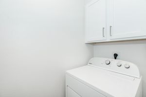 A white washing machine with control dials sits in a small laundry area. Above the machine, there are white cabinets against a plain wall. The space is well-lit and minimalistic, creating a tidy appearance.