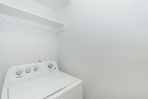 A clean, bright laundry area featuring a white Whirlpool washing machine with control dials. There is a wall-mounted shelf above the machine, and the walls are painted a light color, creating a spacious and uncluttered environment.