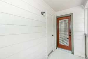 A well-lit entryway featuring white wooden paneling, a glass door with vertical blinds, and a dark-colored door adjacent to it. The area has a clean, modern aesthetic, with natural light illuminating the space.