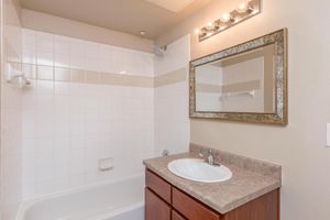 A clean bathroom featuring a bathtub with a showerhead, white tiled walls, and a simple vanity with a sink. A large framed mirror is mounted above the sink, and there is a light fixture with multiple bulbs above the mirror. The decor is neutral and modern, with a subtle beige countertop.