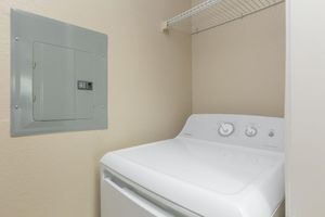 A laundry room featuring a white washing machine next to a circuit breaker panel mounted on the wall. The wall is a neutral color, and there is a shelf above the machine for storage. The setting is clean and organized, suitable for domestic laundry tasks.